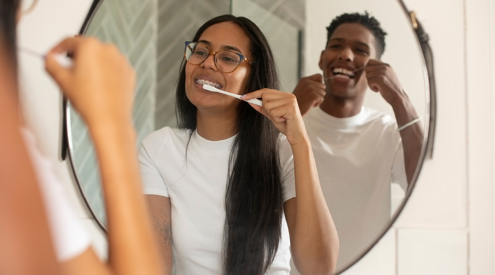 One person brushing their teeth in the mirror and another person flossing in the mirror.